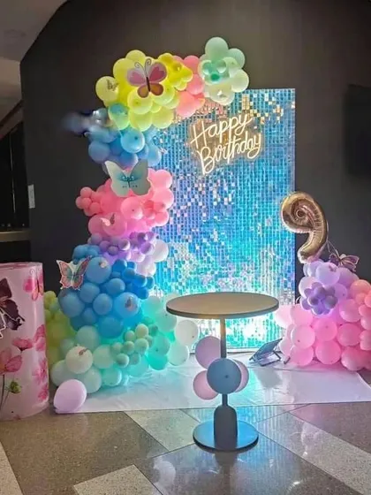 A vibrant butterfly-themed birthday with a blue shimmer wall, a 'Happy Birthday' neon sign, and a rainbow balloon garland.