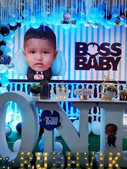 A grand Boss Baby theme for Rudhvik's first birthday celebration. The setup includes a large "ONE" table, multiple character cutouts, and a blue and gold balloon garland against a striped backdrop for a stylish and fun party.