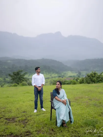 A composed, artistic shot with the bride seated and the groom standing, set against a backdrop of rolling green hills.