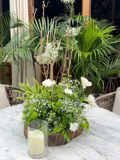 A simple yet elegant centerpiece for a baby shower, featuring fresh white and green florals with dried branches in a wooden base, placed on a marble table.