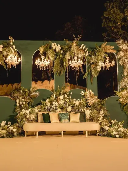 An elegant reception stage set against a deep green backdrop, adorned with white florals and crystal chandeliers. This design combines classic opulence with modern sophistication for a timeless look.
