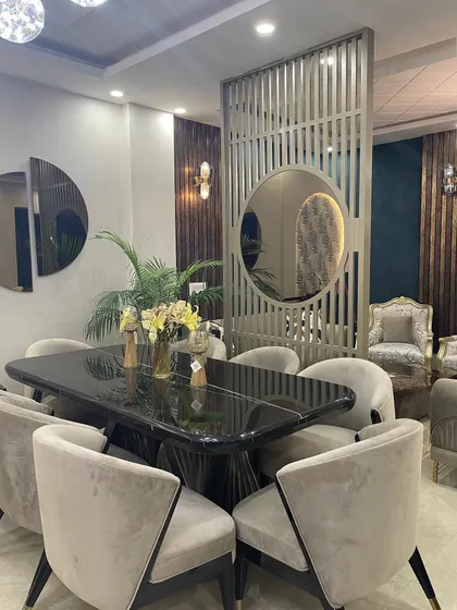 An elegant dining area featuring a large, black marble-top dining table surrounded by plush, beige velvet chairs. A custom slatted wooden partition with a circular mirror adds a contemporary touch and separates the space beautifully.
