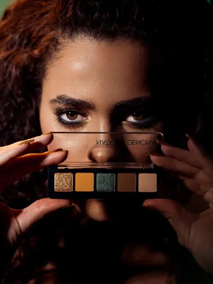 A mysterious and intriguing shot where the model peeks over an eyeshadow palette. The dramatic, low-key lighting casts deep shadows, drawing all the attention to her intense eyes and the product. It’s a perfect image for a campaign that wants to create suspense and allure.