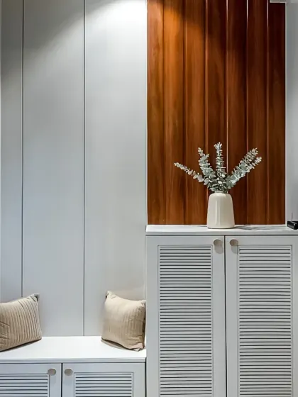 A clean and simple entryway design featuring a white shoe cabinet with louvered doors for ventilation. The unit is paired with a matching bench and a contrasting wood-paneled accent wall.