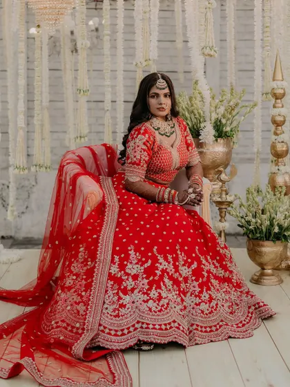 A regal portrait of the bride seated, allowing her magnificent red lehenga to be displayed in its full glory. The composition and decor create a sense of royal elegance.