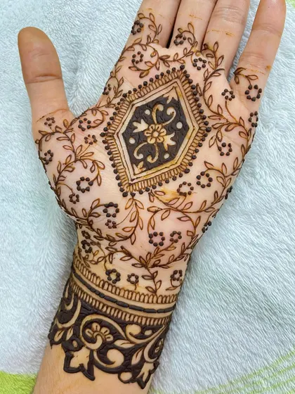 A beautiful party henna design with a central geometric floral motif. The palm is surrounded by delicate leafy vines, and the wrist has a bold, dark cuff.