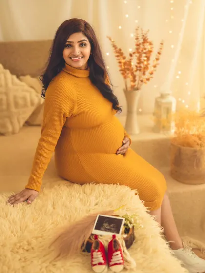 A cozy and warm portrait of a mom-to-be in a mustard yellow knit dress, seated in our boho studio with her baby's first shoes and sonogram.