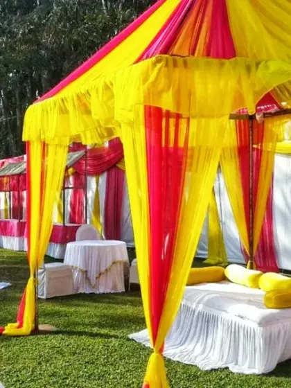 Colorful canopies (shamianas) set up on a lawn for a daytime wedding event. These tents provide seating areas for guests and are decorated in bright yellow and pink fabrics.