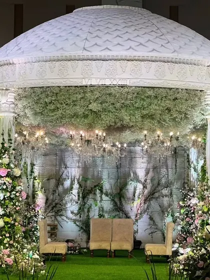 The main mandap structure, a white dome set against a backdrop of green walls and floral trees, creating a serene and picturesque setting for the ceremony.