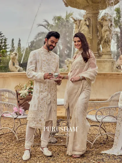 A moment of shared joy. The groom's gold-embroidered sherwani and the bride's simple saree create a beautiful contrast, both unified by a palette of ivory and gold.