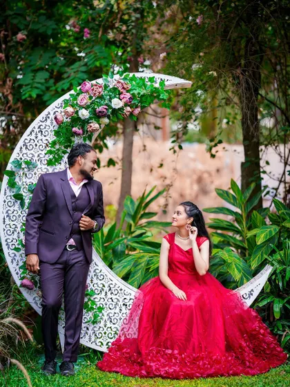 A whimsical pre-wedding portrait featuring a couple posing with a large, ornate crescent moon prop decorated with flowers.