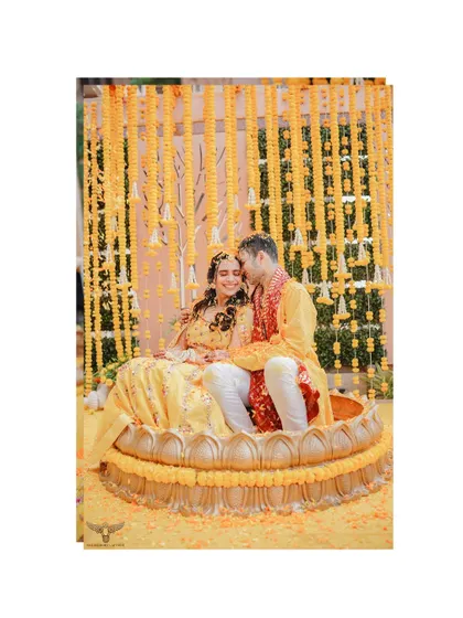 A sweet, intimate moment during the Haldi. The couple is seated in a giant lotus-shaped vessel, sharing a quiet, loving glance amidst the vibrant yellow decor.
