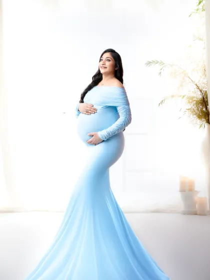 A classic, light-filled portrait. The expectant mother looks radiant in a floor-length blue gown, posed against a bright window for a timeless and elegant feel.