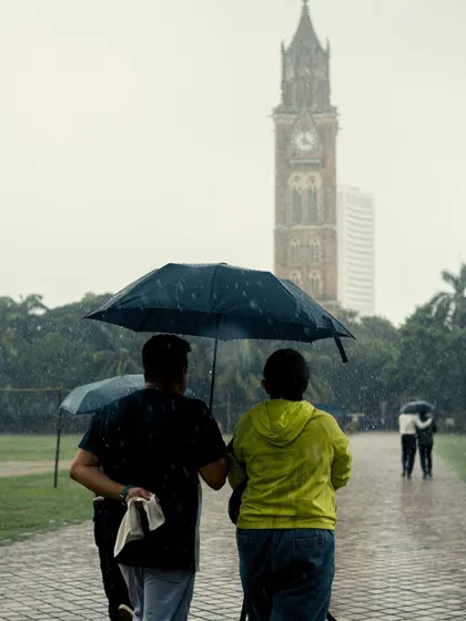 Two men walk together under an umbrella at Oval Maidan. It's a simple, candid shot of companionship, with the Rajabai Clock Tower providing a classic Mumbai backdrop.