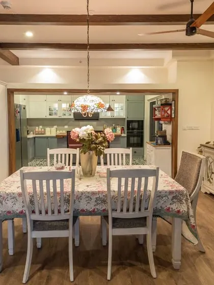 A charming dining room that blends classic and rustic elements. The distressed white furniture and floral tablecloth create a vintage feel, while the exposed wooden beams add an industrial touch.