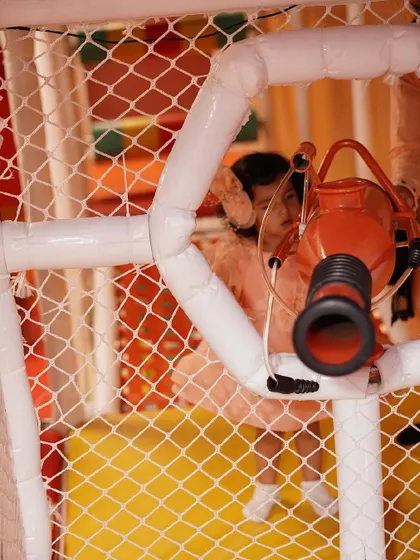 A child peeks through the netting of our intricate play structure, ready to launch a soft ball from a cannon.