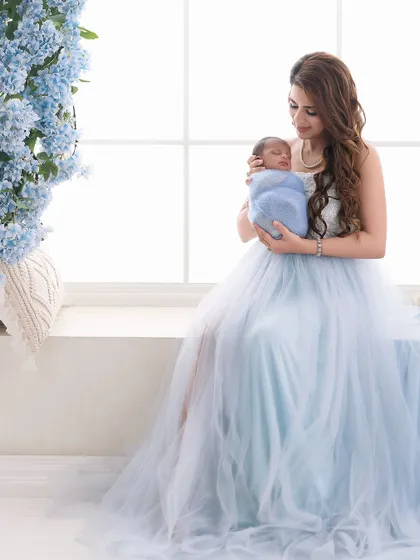 A full-length portrait of a mother in a flowing blue gown holding her swaddled newborn. The composition uses natural light and a hint of floral decor to create an elegant and dreamy image.