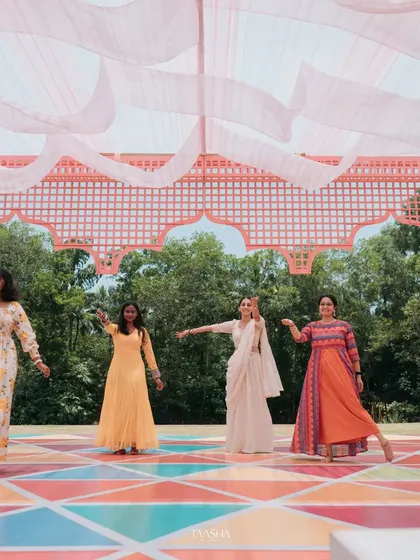 A colorful dance performance at a daytime Mehendi event. The stage features a vibrant, multi-colored floor and a pink canopy, creating a joyful and high-energy setting.