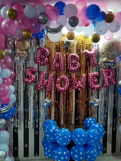 A classic baby shower decoration with a silver shimmer curtain and "BABY SHOWER" foil balloons. The setup is framed by a large balloon arch using pink, blue, white, and gold balloons.