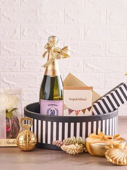 A celebratory hamper perfect for an engagement or wedding announcement. It includes a bottle of champagne, a scented sachet, and other small gifts, all packed in a chic black-and-white striped hat box.
