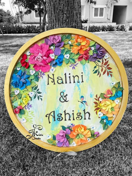 A stunning 14-inch circular nameplate for "Nalini & Ashish," framed with a vibrant garden of multi-colored sculpted flowers. The black and white background highlights the colorful floral art.