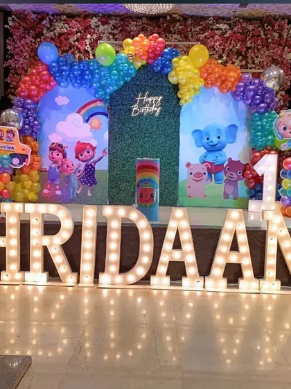 A large-scale Cocomelon themed stage decoration for a first birthday. The setup includes a huge backdrop with characters, a colorful balloon arch, and large light-up marquee letters spelling out the name "HRIDAAN".