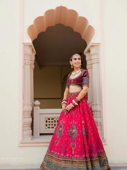 A classic bridal portrait framed by a traditional Rajasthani archway. The vibrant pink of her lehenga stands out against the neutral stone.