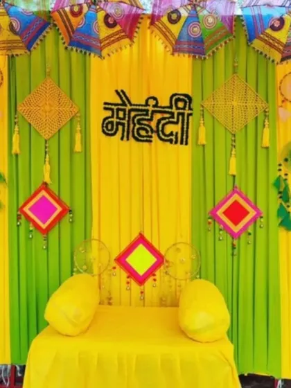 A playful and colorful Mehendi backdrop with bright yellow and green drapes. The decor is enhanced with Rajasthani umbrellas, dreamcatchers, and a 'Mehendi' sign made of flowers.