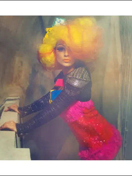An edgy fashion editorial with a hazy, dreamlike quality. The vibrant yellow wig and colorful outfit pop against the gritty background, creating a striking contrast.