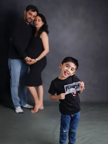 A creative pregnancy announcement photo. The older brother stands in the foreground proudly holding the ultrasound picture, while his parents embrace in the background, creating a fun and memorable family moment.