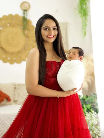 A joyful mother in a red gown holding her sleeping newborn. Her bright smile perfectly captures the happiness of motherhood.