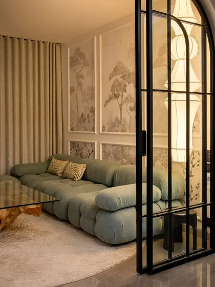 An intimate oasis of calm. This cozy nook features a plush, curved sofa set against a wall with nature-inspired art panels. The arched glass door adds a classic touch, separating the space while maintaining a visual connection.