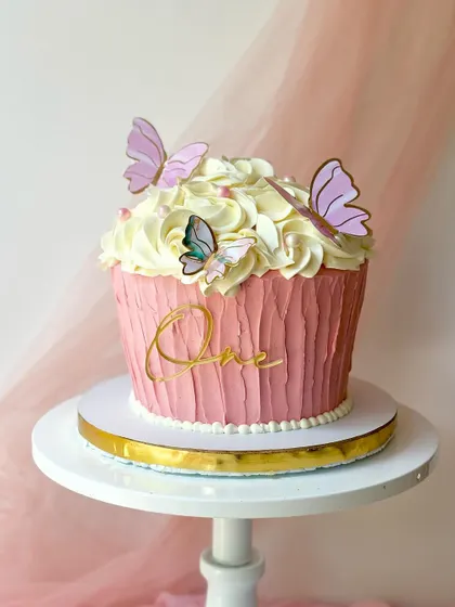 A giant cupcake cake for a first birthday, with a textured pink base and fluffy white buttercream frosting, adorned with butterflies.