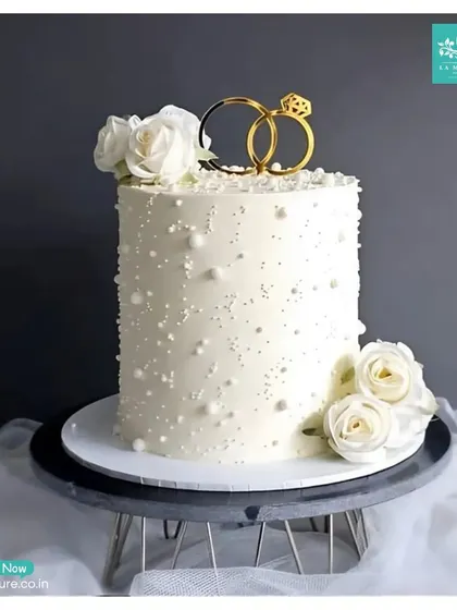 Simplicity at its finest. This tall, single-tier engagement cake is decorated with delicate sugar pearls and white roses, topped with an elegant intertwined rings topper, making it a perfect choice for a chic and modern celebration.