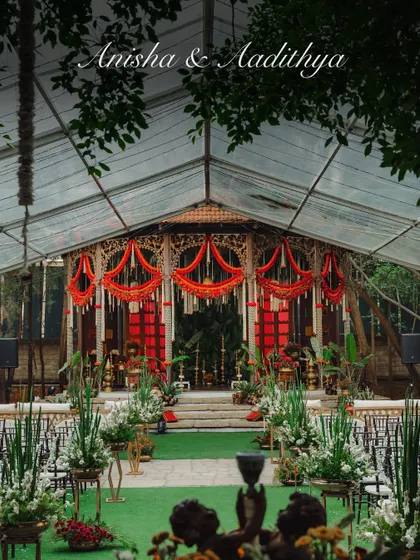 A majestic wedding setup under a transparent German tent, ensuring weather is never a concern. The grand mandap, with its bold red and orange floral decor, stands as a vibrant centerpiece, ready for a traditional South Indian wedding.