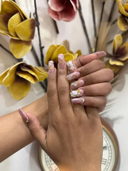 An exquisite set of long, square-tipped bridal nails. This design features a soft pink base with intricate 3D white flowers, gold charms, and sparkling rhinestones for a truly unforgettable look.