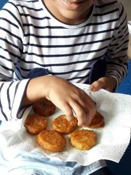 A child selects a warm aloo tikki from the plate. The sensory experience of touching, smelling, and tasting the food is an integral part of the learning process.