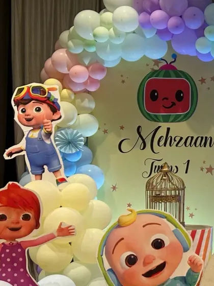 A detailed view of a Cocomelon party backdrop, featuring character cutouts, a personalized name sign, and a pastel balloon arch.
