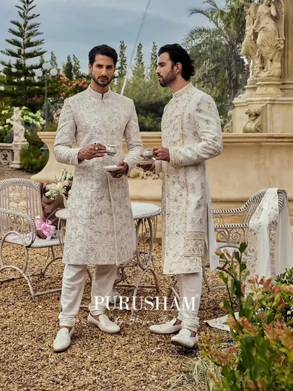 In the heart of elegance, two gentlemen savor a quiet moment. I designed these coordinated ivory sherwanis, one with geometric embroidery and the other with floral, to showcase individual style within a shared aesthetic.