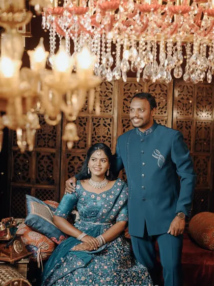 A formal portrait of a couple on a royal-themed set, her in a blue lehenga and him in a matching bandhgala.