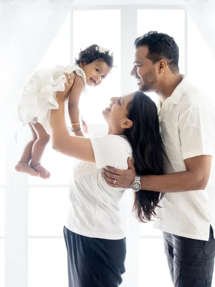 A joyful mother lifts her daughter in the air as the father looks on lovingly. A perfect picture of family happiness.