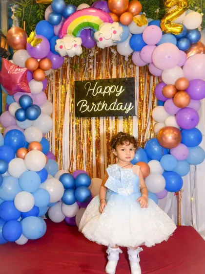 A full-length shot of the birthday girl in her pretty blue and white dress, standing in front of her rainbow-themed birthday decorations.