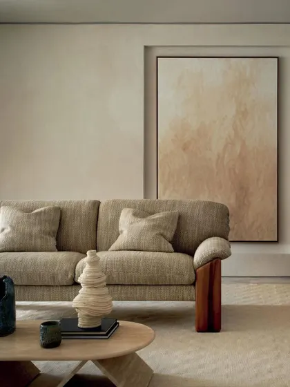 A full view of a sofa upholstered in the "Selma" fabric from the Prestige II collection, showcasing its graceful, flowing weave. Set in a minimalist, neutral space, the sofa's texture and solid wood arm details become the quiet focal point.
