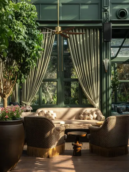 A sun-drenched seating area in the Lord Elgin Glasshouse. The design layers rich, patterned fabrics, fringed armchairs, and warm brass accents against the deep green of the structure to create a sense of exotic escapism.