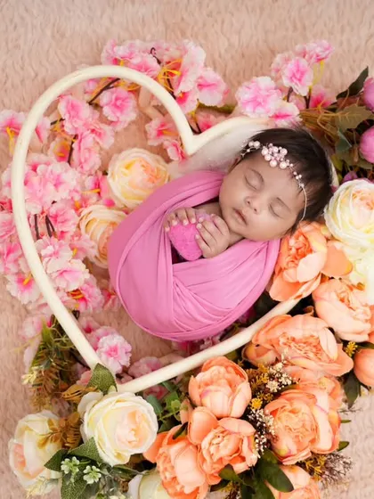 A newborn wrapped in a soft pink swaddle sleeps inside a heart-shaped frame, surrounded by an arrangement of beautiful pink and peach flowers. This is a perfect way to capture the love for your new arrival.