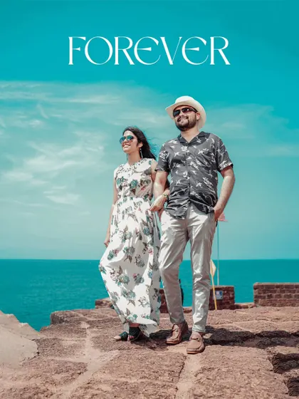 A beautiful, wide-angle shot of the couple on the coast of Goa, with the bright blue sky and sea creating a stunning backdrop for their "Forever" moment.