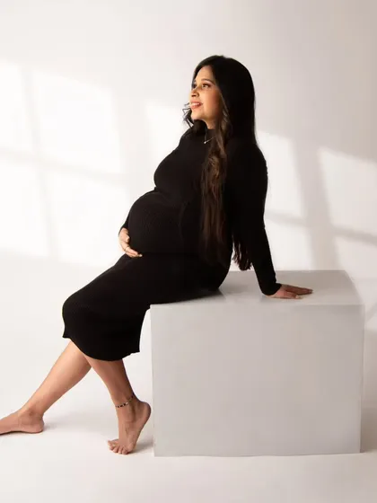 A modern and chic portrait. The mom-to-be sits on a white block, her black dress creating a clean silhouette against the bright background.