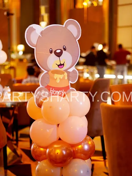 A close-up of a cute teddy bear prop holding a honey pot, placed on a column of orange and gold balloons.