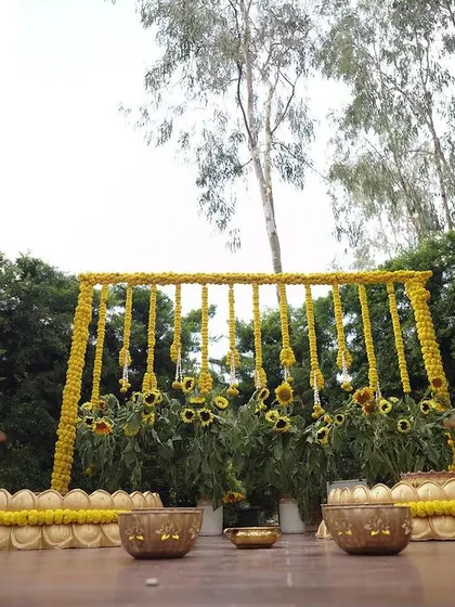 A low-angle view of our nature-inspired Haldi decor, showing the traditional brass urlis and the sunflower backdrop, all set against a canopy of trees.