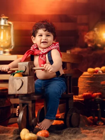 This little cowboy is full of joy, sitting on his wooden tricycle in a barn-themed setup with pumpkins and hay.